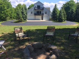 View of parking area and fire pit at back Torch Lake lodge. Includes outdoor seating. Yard is available for games. Also shows ample area for parking of toys.